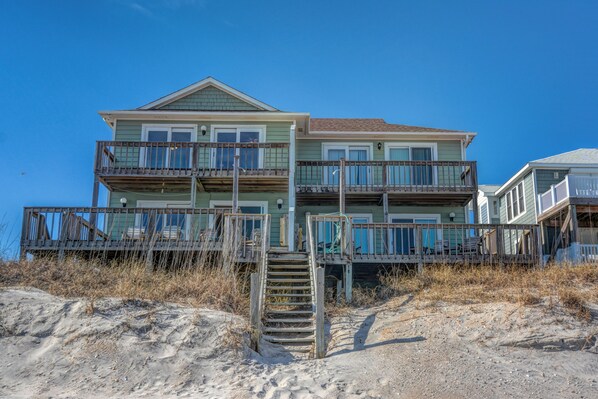 Una casa frente al mar de varios pisos con una terraza de madera y una escalera que conduce a la entrada.