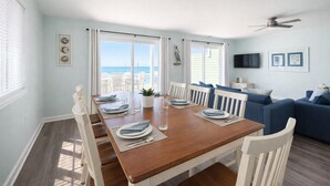 A dining area with a wooden table set for four, white chairs, and a view of the ocean through large windows.