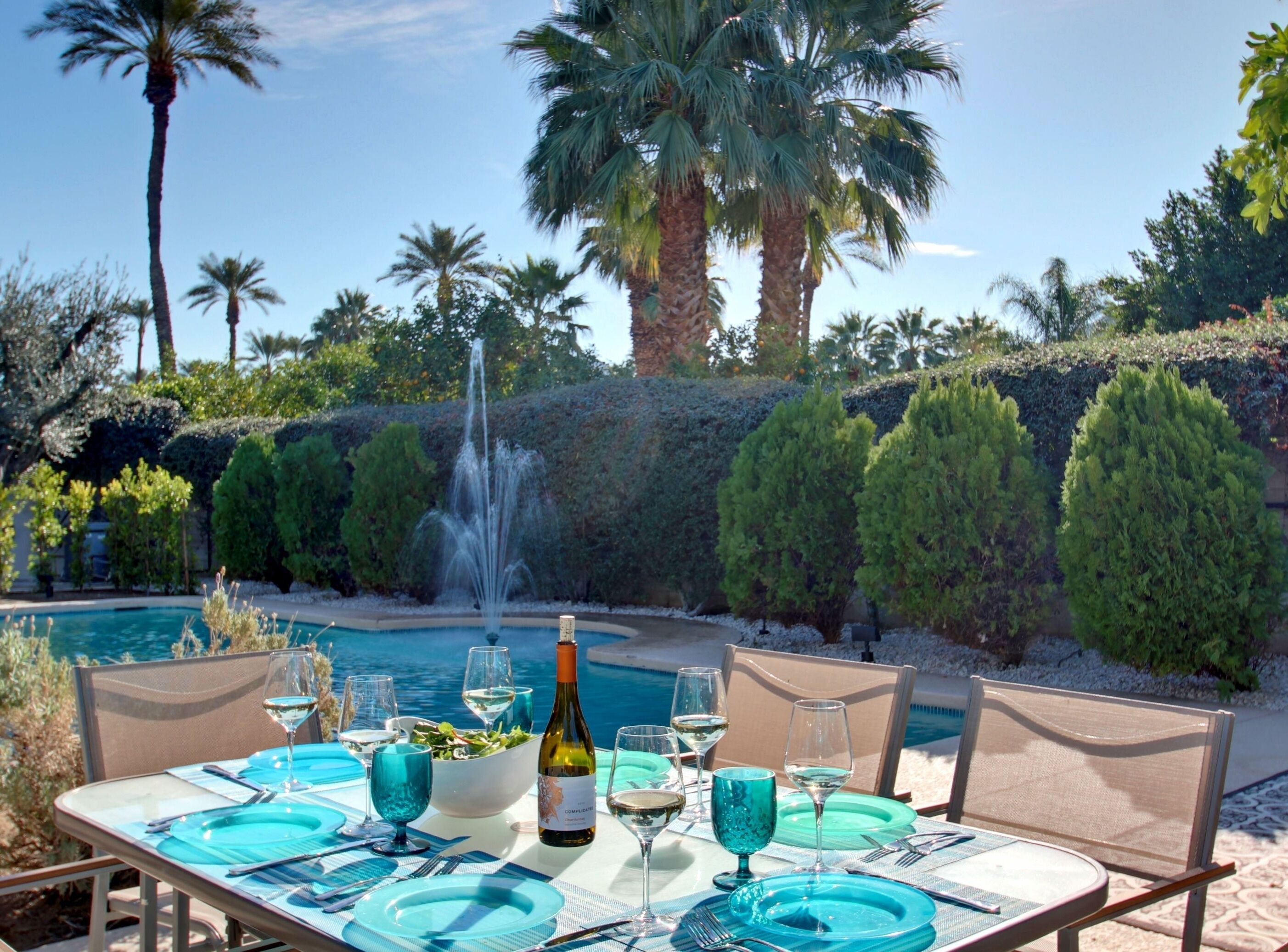 A dining table set with blue tableware and a bottle of wine, overlooking a pool and palm trees.