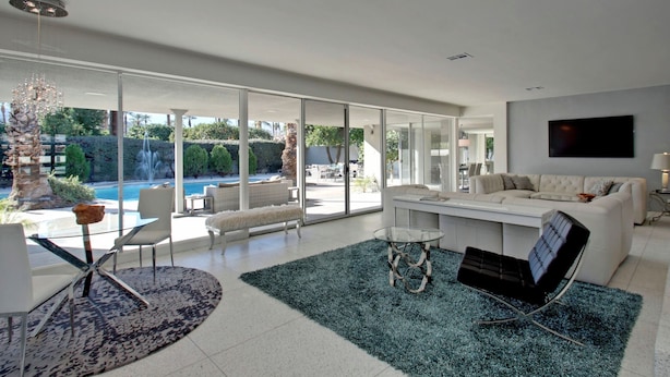 Uma sala de estar moderna com uma grande porta de vidro que dá para uma área de piscina, um sofá branco, uma mesa de centro de vidro e uma TV de tela plana na parede.