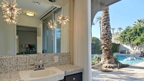 Casa de banho com um espelho grande, lavatório e vista para a piscina e uma palmeira lá fora.