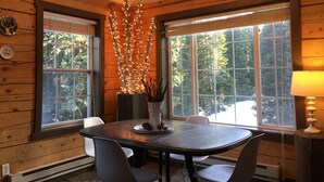 A wooden cabin interior with a dining table, chairs, a chandelier, and large windows with a forest view.