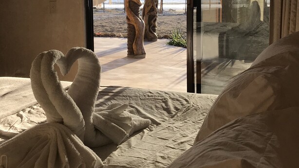 A room with a bed, a heart-shaped towel arrangement, a giraffe sculpture, and a view of the beach and ocean through a glass door.