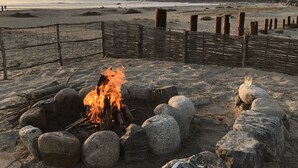 A beach scene with a fire pit surrounded by rocks, a fence, and a distant view of a hill with buildings.