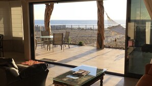 A spacious living room with a large glass door leading to a beach view. Inside, there is a sofa, a coffee table with magazines, and a ceiling fan.