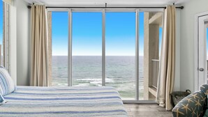 Ein Schlafzimmer mit einem großen Bett, Meerblick durch raumhohe Fenster und ein Balkon mit einem Stuhl.