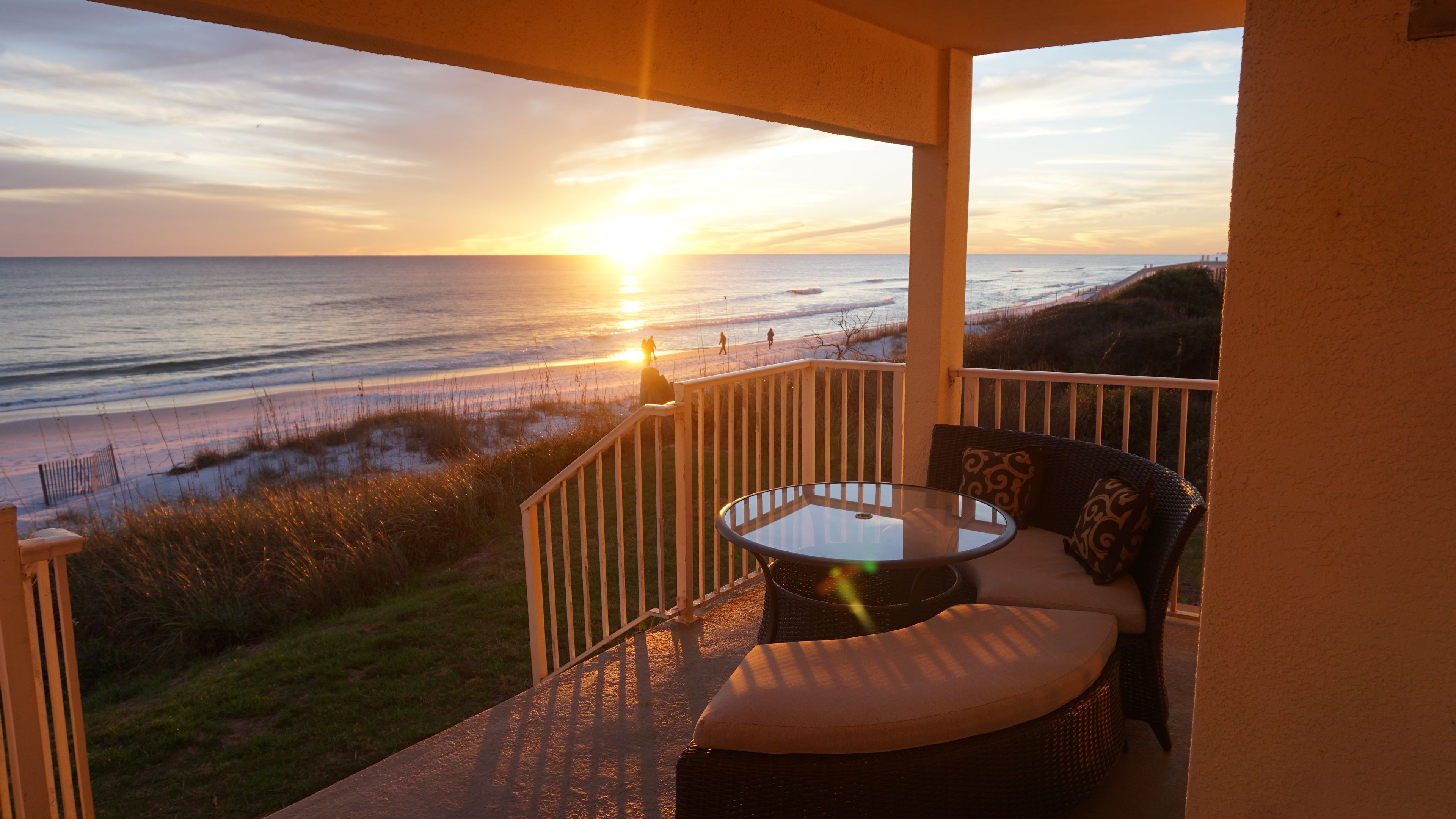 A balcony with a glass table and wicker furniture overlooking a beach at sunset.