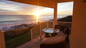 A balcony with a glass table and wicker furniture overlooking a beach at sunset.