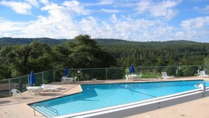 A rectangular swimming pool with a clear blue water, surrounded by white lounge chairs and blue umbrellas, set against a backdrop of a forested hill under a partly cloudy sky. - Woodsy Hideaway~Yosemite views, your getaway starts here, it's a Guest Favorite! (Groveland)