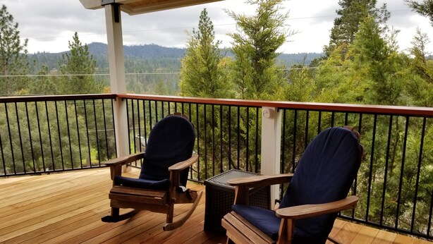 A wooden deck with two rocking chairs and a small table, overlooking a forested landscape. - Woodsy Hideaway~Yosemite views, your getaway starts here, it's a Guest Favorite! (Groveland)