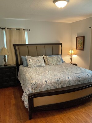 A bedroom with a bed, a nightstand, a lamp, a window with curtains, and a wall with a framed picture.