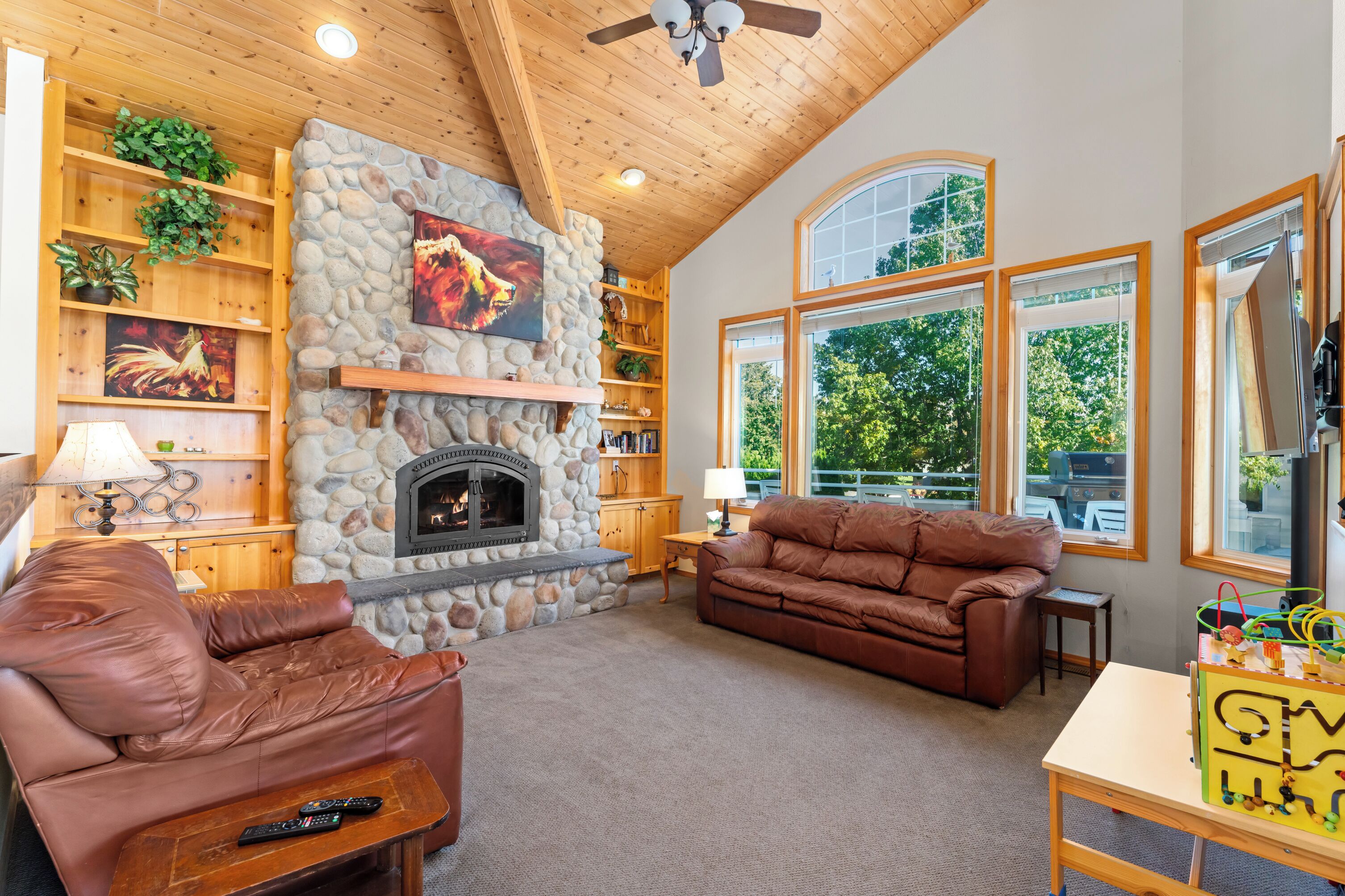 A cozy living room with a stone fireplace, leather sofas, wooden shelves, and large windows.