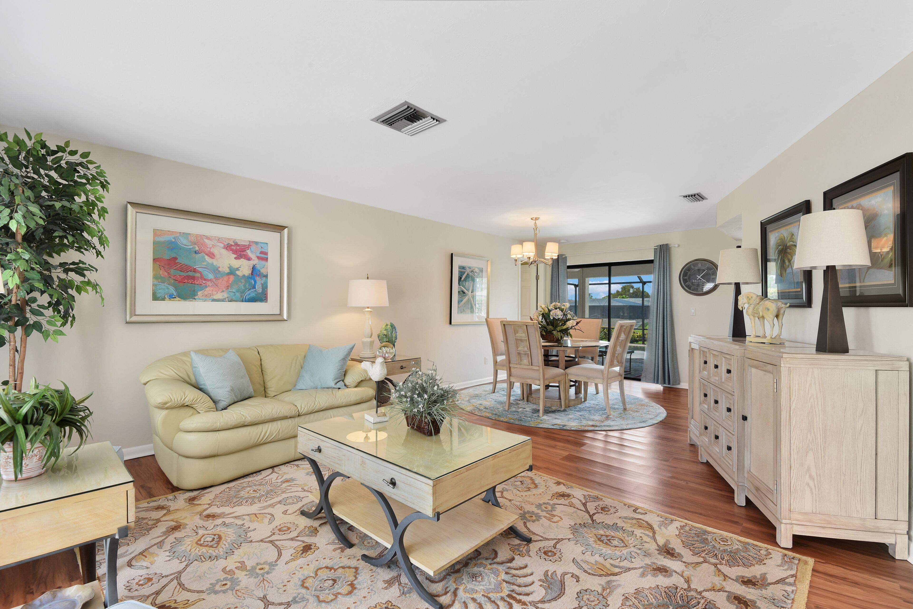A living room with a beige sofa, a glass coffee table, a dining area with a table and chairs, and a light-colored cabinet.