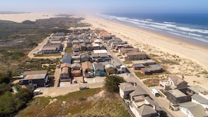 An aerial view of a coastal residential area with houses along the beach, clear skies, and a visible road.