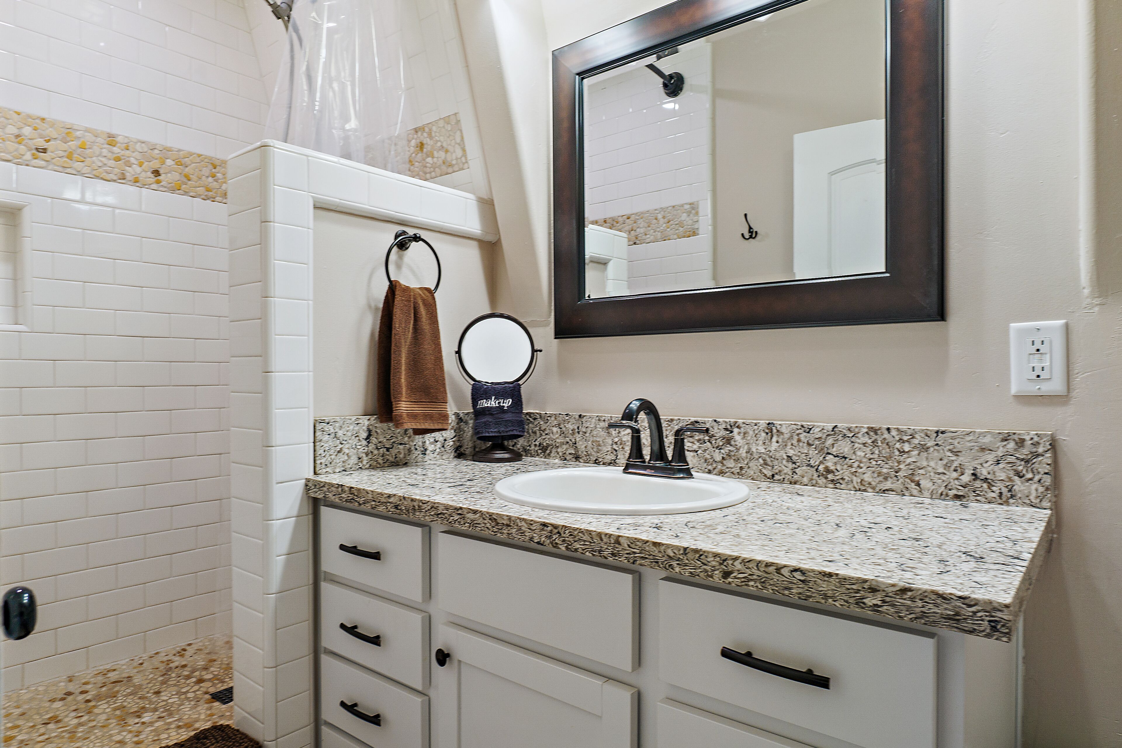Une salle de bain avec des murs carrelés de carreaux blancs, un comptoir en granit, un lavabo avec un robinet, un miroir et un porte-serviettes avec une serviette marron.