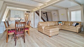 A spacious living area with a large L-shaped leather sofa, wooden dining table with chairs, and a kitchen in the background.