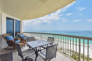 Un balcon avec des fauteuils en osier et une table, donnant sur une plage parsemée de parasols et fréquentée par des gens.