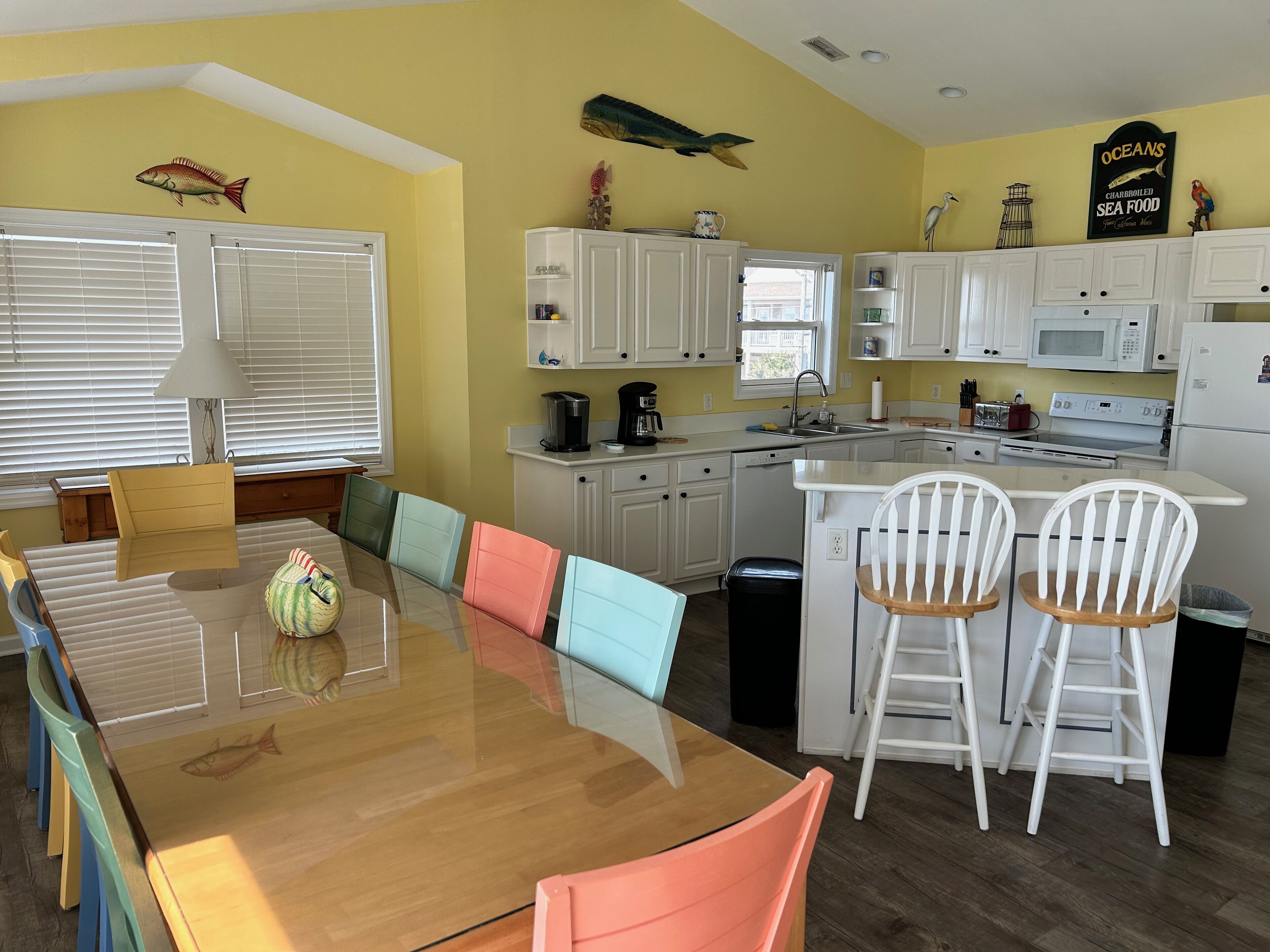 Une cuisine avec des meubles blancs, une table à manger en bois entourée de chaises colorées et un îlot de cuisine avec deux chaises blanches.