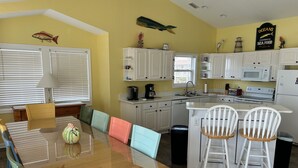 Une cuisine avec des meubles blancs, une table à manger en bois entourée de chaises colorées et un îlot de cuisine avec deux chaises blanches.