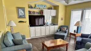 Un salon comprenant un canapé, un fauteuil, une table basse, ainsi qu’un coin cuisine avec des placards et un téléviseur.