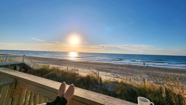 Une terrasse en bois donnant sur une plage, d’où l’on a une vue dégagée sur l’océan et sur le soleil se couchant à l’horizon.