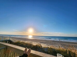 Een houten terras met uitzicht op een strand, waar de oceaan zichtbaar is en de zon in de verte ondergaat.