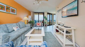 Ein Wohnzimmer mit Sofa, Couchtisch und Fernseher. Es gibt Pflanzen, einen Deckenventilator und einen Balkon mit Blick.