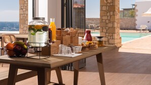 Frühstücksaufstellung mit Obstkorb, Wasserspender und verschiedenen Frühstücksleckereien auf einem Holztisch am Pool.