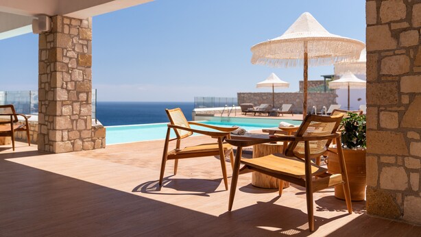 Ein sonnendurchfluteter Außenbereich mit Holzmöbeln und weißen Sonnenschirmen, mit Blick auf einen Pool und das Meer.