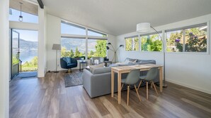 Un salon moderne comprenant un canapé, une table à manger et des chaises, de grandes fenêtres offrant une vue sur un lac et des montagnes, ainsi qu’une lampe.