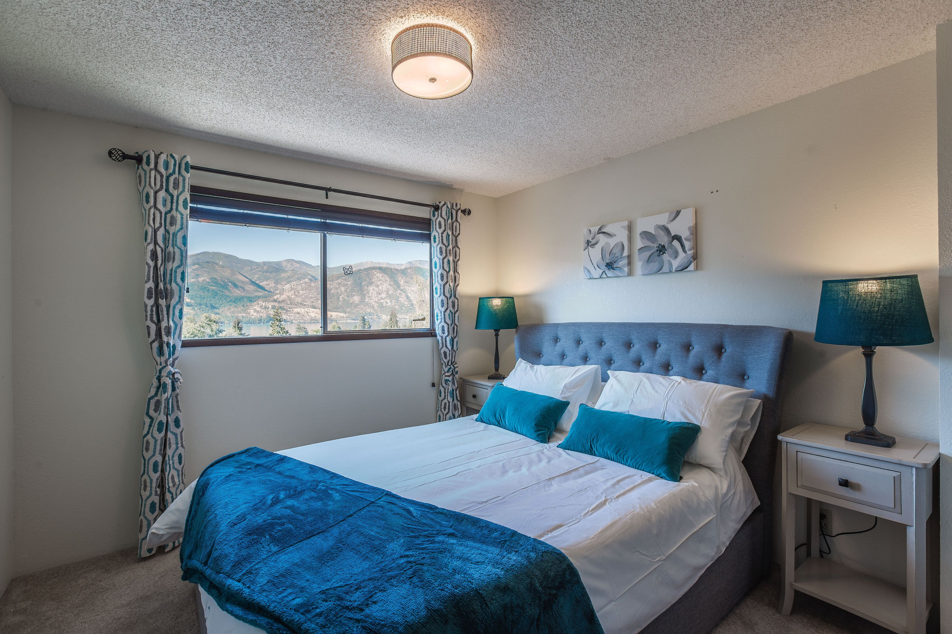 A bedroom with a bed, two bedside tables, a window with curtains, a ceiling light, and a view of mountains.