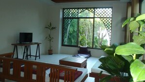 Una habitación con televisión sobre un mueble bajo, un sofá, una ventana que ofrece vistas a zonas verdes y una planta grande en maceta.