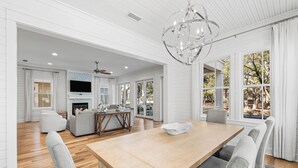 A spacious living room with a wooden dining table, grey upholstered chairs, a fireplace, and a large TV mounted on the wall. - Stunning WaterColor Retreat: Bikes, WaterColor Beach Club Access, Carriage House, Pure Relaxation! (Santa Rosa Beach)