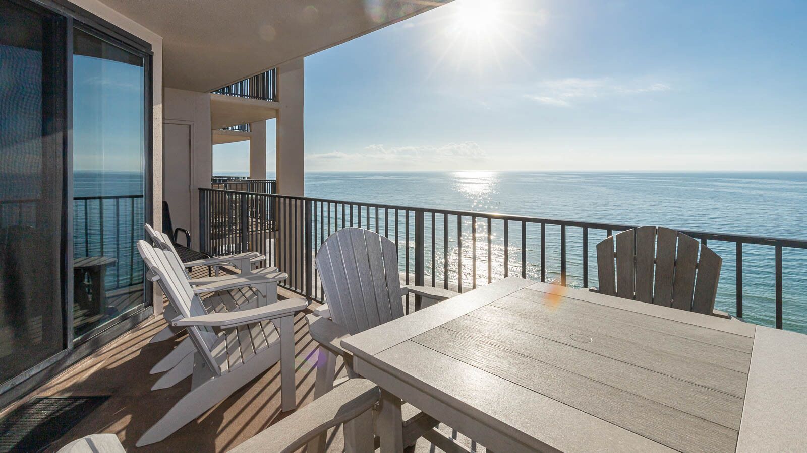 A balcony with a view of the ocean, a table, and two Adirondack chairs.