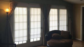 A living room with a sofa, ottoman, and a lamp. There are large windows with curtains, a ceiling fan, and a door. - Pelican condo in Chimney Creek (Tybee Island)
