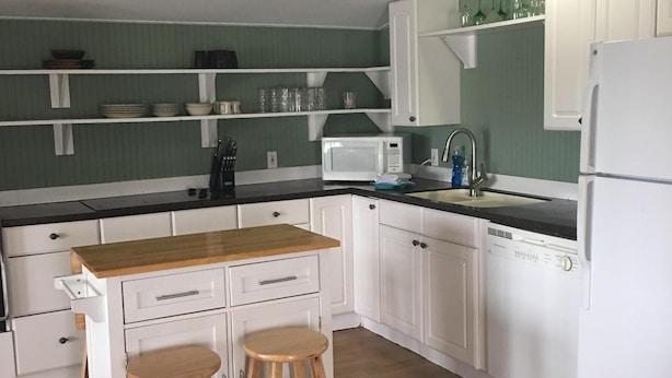 A compact kitchen with white cabinets, a microwave, a sink, and a refrigerator. There are two wooden stools at the counter. - Pelican condo in Chimney Creek (Tybee Island)