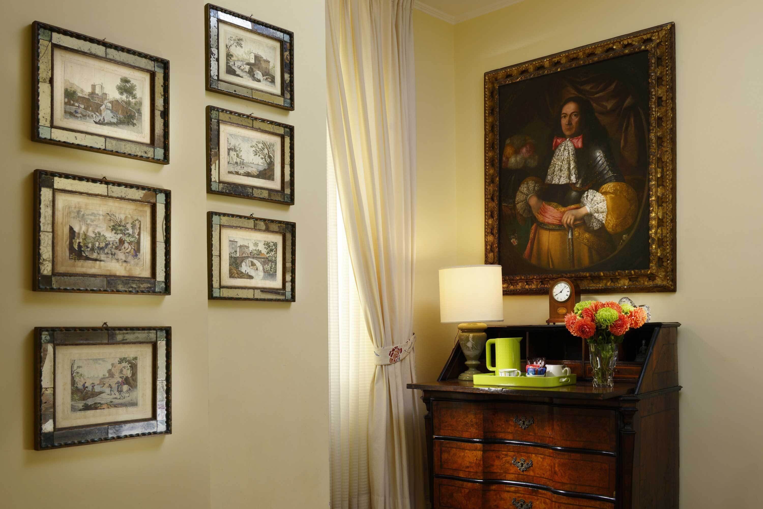 A room with a wall decorated with framed pictures, a large painting, a wooden dresser with drawers, a green mug, and a vase with flowers.