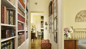 A narrow hallway with bookshelves on one side and a bed with a headboard on the other. There is a chair in the hallway and a dining table in the background. - Close to Navona Square, Exclusive cozy Apt AC, WiFi. Perfect base for touring (Roma)