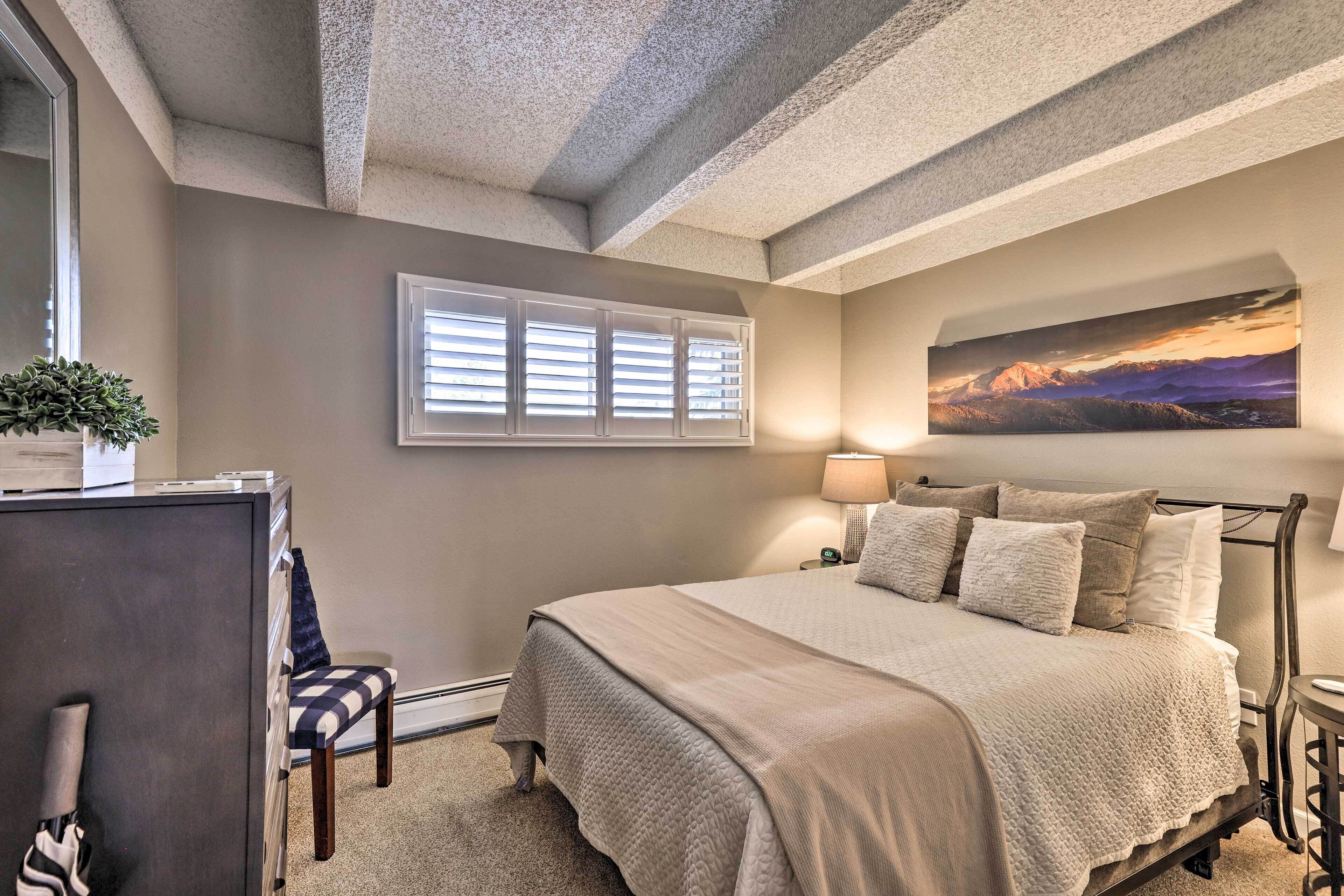 A bedroom with a large bed, a window with blinds, a painting on the wall, a desk, and a chair.