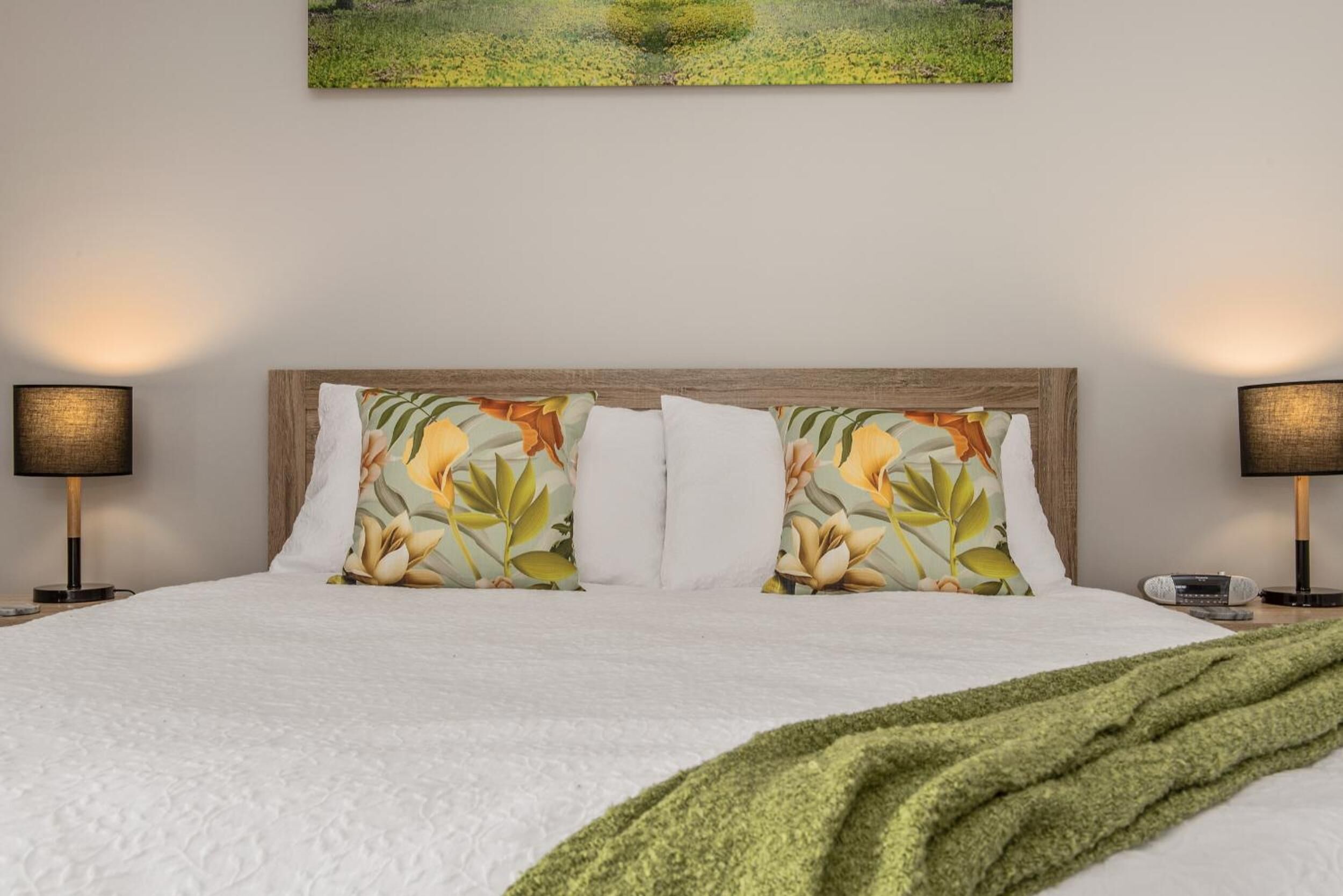 Ein ordentlich bezogenes Bett mit weißen Leinentüchern, zwei Kissen mit Blumenmuster und einer grünen Tagesdecke. An jeder Seite des Bettes stehen zwei Nachttischlampen.