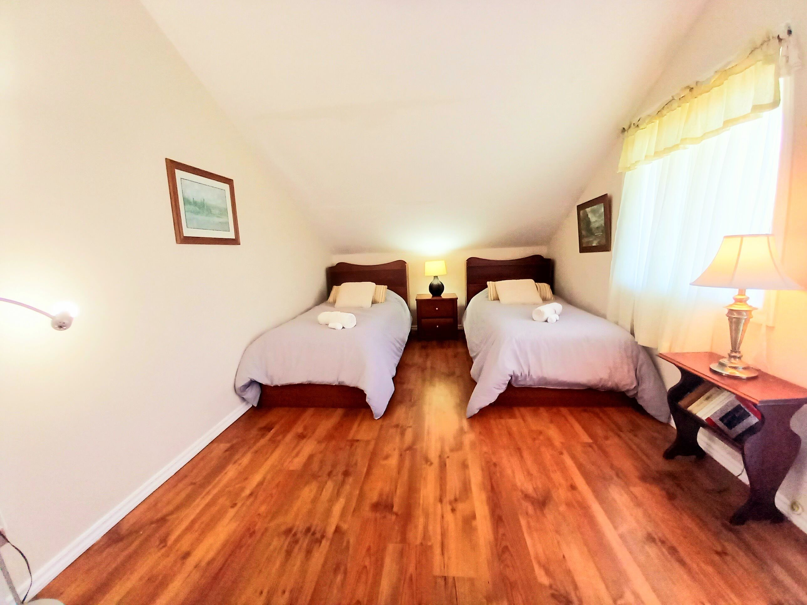 A room with two beds, wooden flooring, a nightstand with a lamp, and a framed picture on the wall.