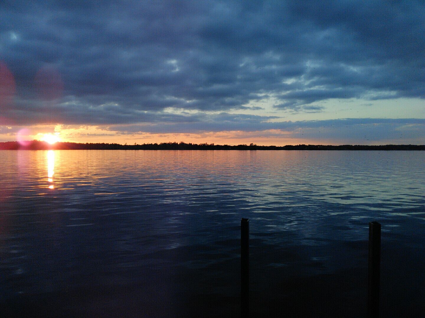 A calm body of water reflecting the sunset with a distant shoreline and overcast sky.