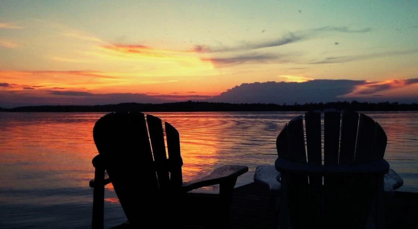 Two Adirondack chairs facing a sunset over a body of water.