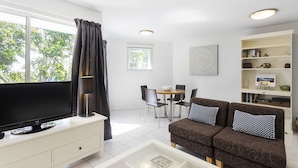 A modern living room with a flat-screen TV, a sofa with patterned cushions, a bookshelf, and a dining area with a table and chairs. - Byron Getaway at Eco Beach (Byron Bay)
