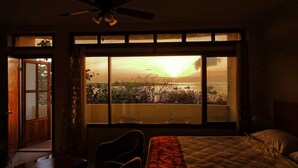 A room with a bed, a desk, and a large window with a view of a sunset over a body of water.