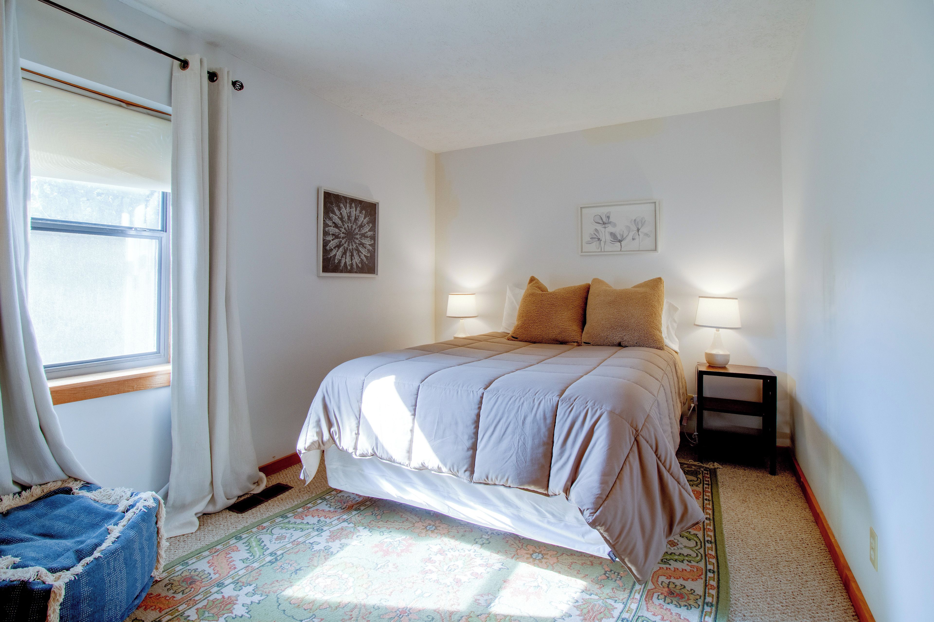 Una camera da letto con un letto, un comodino, una lampada, una finestra con tende e una parete decorata da due quadri.