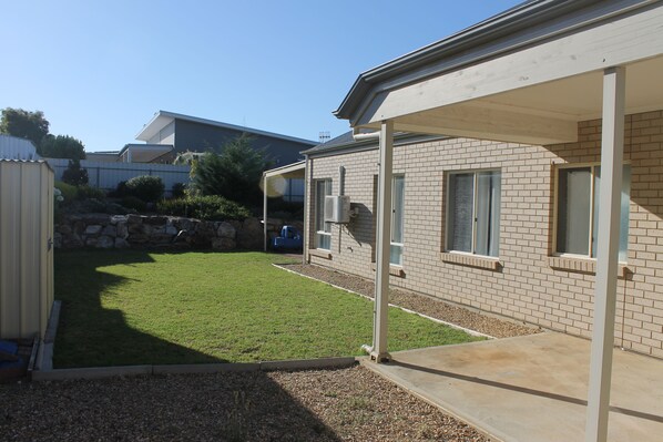 A residential backyard with a lawn, a covered patio, and a brick house. - Pebbles Cottage in Normanville (Normanville)