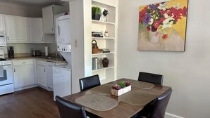 Una cocina moderna con armarios blancos, una zona de comedor con una mesa de madera y sillas negras, y un cuadro de flores en la pared.
