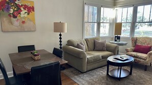 Una sala de estar con un sofá color beige, una mesa redonda para café, una mesa de comedor con sillas y un sillón estampado con motivos florales.