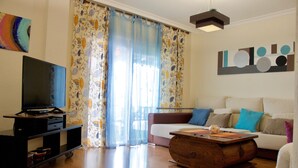 Un salon avec un téléviseur à écran plat posé sur un meuble, une table basse en bois, un canapé avec des coussins et une grande fenêtre ornée de rideaux à motifs floraux.